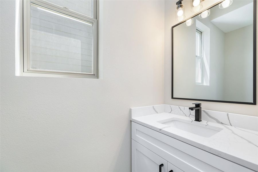The beautiful contrast of white and black seen in the kitchen carries into the bathrooms, where a sleek white countertop is complemented by bold black fixtures. A large framed mirror and ample natural light from a nearby window create a bright and inviting atmosphere.