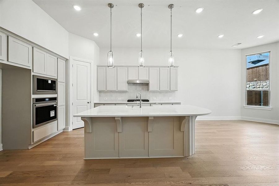 Kitchen with decorative backsplash, light wood-style floors, pendant lighting, a breakfast bar area, and a kitchen island with sink Kitchen with decorative backsplash, light wood-style floors, pendant lighting, a breakfast bar area, and a kitchen island with sink