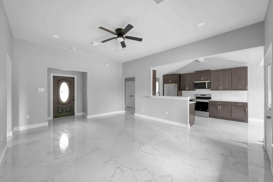 This spacious living area features sleek marble floors, a ceiling fan, and an open layout leading to a modern kitchen with dark cabinetry and stainless steel appliances. The room is well-lit with natural light, creating a bright and inviting atmosphere.