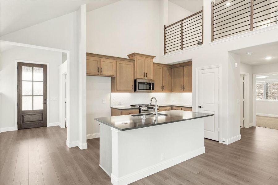 Open kitchen with center island, dark countertops, tan cabinets, stainless appliances, and two-story ceiling with upper railing. Open kitchen with center island, dark countertops, tan cabinets, stainless appliances, and two-story ceiling with upper railing.