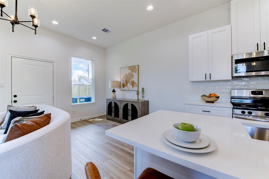 View from kitchen island toward living area and rear exit, showing hardwood floors, recessed lighting, chandelier, white shaker upper cabinets, stainless appliances, and subway tile backsplash.