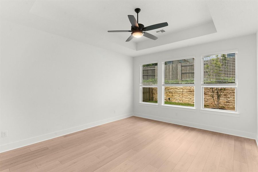 Spacious room featuring light wood-finish flooring, a tray ceiling, and a contemporary ceiling fan with integrated lighting Spacious room featuring light wood-finish flooring, a tray ceiling, and a contemporary ceiling fan with integrated lighting