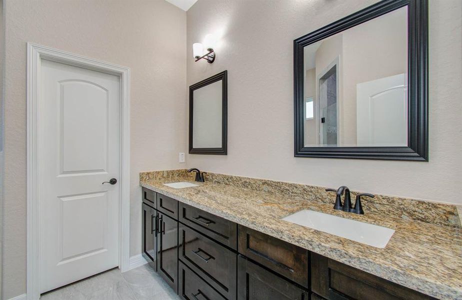 This is a modern bathroom featuring a granite countertop with an undermount sink, dark cabinetry, a framed mirror, and elegant wall sconces. A single white door suggests privacy and convenience.