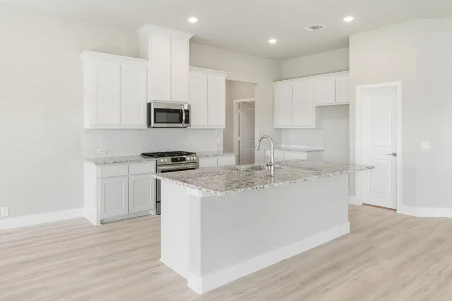 Kitchen with white cabinets, granite island with sink, stainless steel appliances, and light wood floors