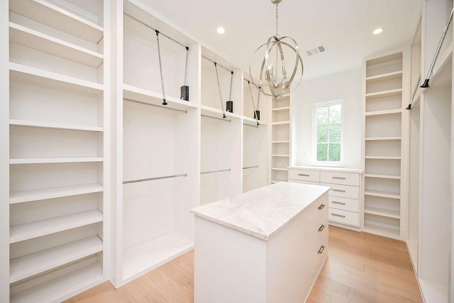 Primary Walk-in closet-spacious and Luxurious with Over-Under Custom Cabinetry. Island and Chandelier Accent. featuring open shelves, hanging rods, and a central island with drawers.
