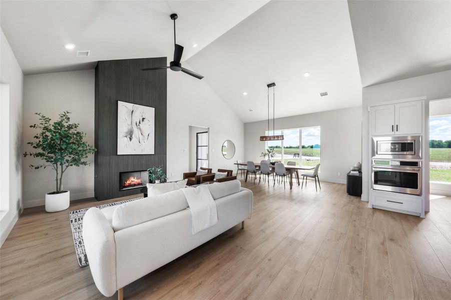Living area with light wood-style flooring, a large fireplace, ceiling fan, plenty of natural light, and high vaulted ceiling Living area with light wood-style flooring, a large fireplace, ceiling fan, plenty of natural light, and high vaulted ceiling