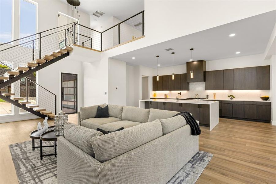Open-concept living space featuring a floating staircase with cable railing, wood-finish flooring, a kitchen with dark cabinetry, a center island, and integrated lighting