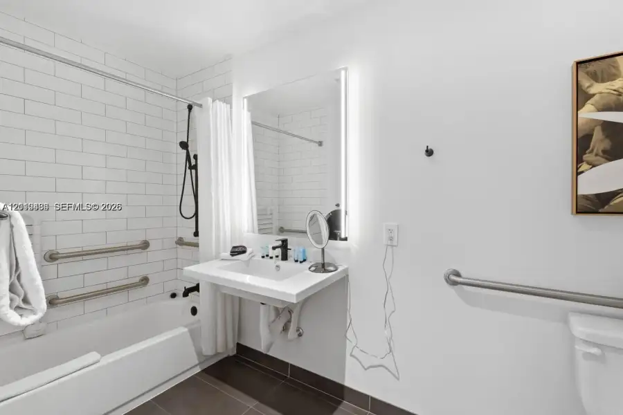 Wall-mounted floating vanity with integrated sink, rectangular illuminated mirror, white subway-tiled shower surround, matte black plumbing fixtures, and installed safety grab bars Wall-mounted floating vanity with integrated sink, rectangular illuminated mirror, white subway-tiled shower surround, matte black plumbing fixtures, and installed safety grab bars