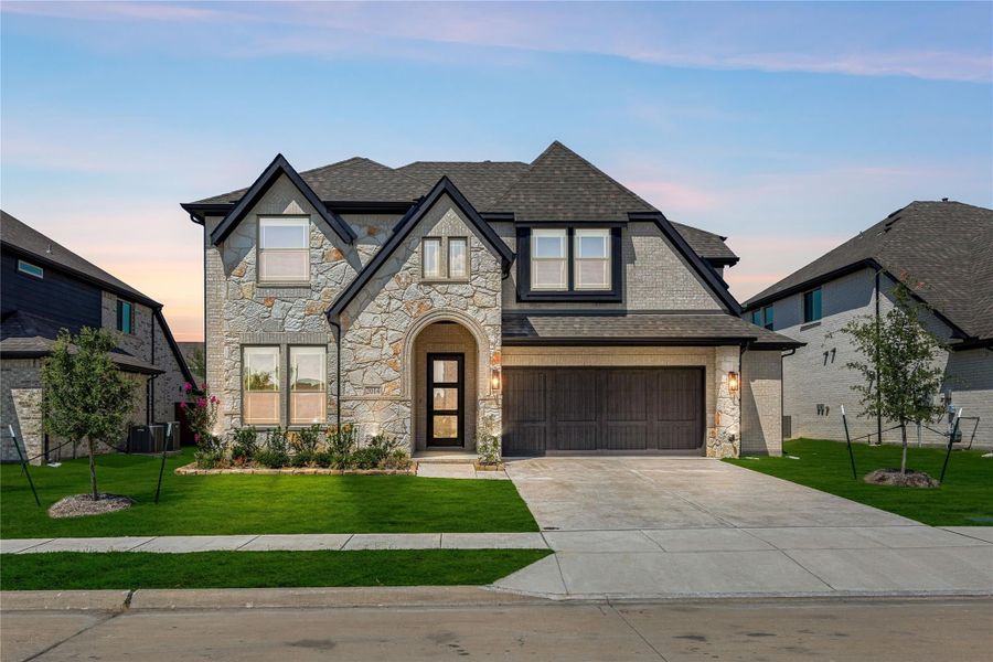 Two-story home with stone and brick exterior, arched entry, two-car garage, and manicured front lawn at dusk