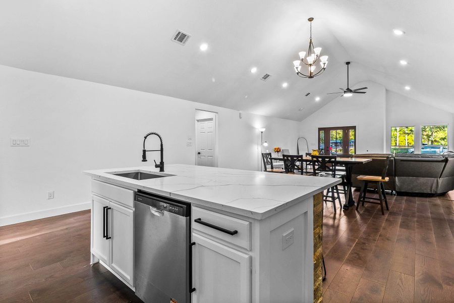 This photo showcases an open-concept living area with a vaulted ceiling, modern kitchen island, and hardwood floors. The space includes a dining area and a cozy seating arrangement, accented by large windows and stylish lighting fixtures.