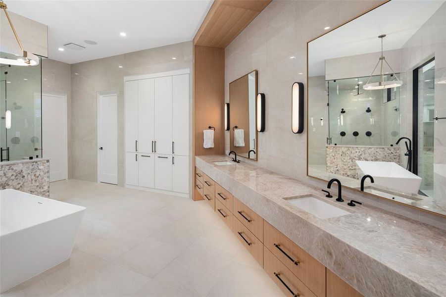 Luxurious primary bathroom featuring a spa-like design with an expansive walk-in shower, an expansive vanity with Taj Mahal quartzite counters, dual sinks, custom cabinetry, and designer lighting. Elegant finishes and natural textures create a serene retreat, while built-in linen cabinetry with a hidden hamper adds both convenience and thoughtful functionality.