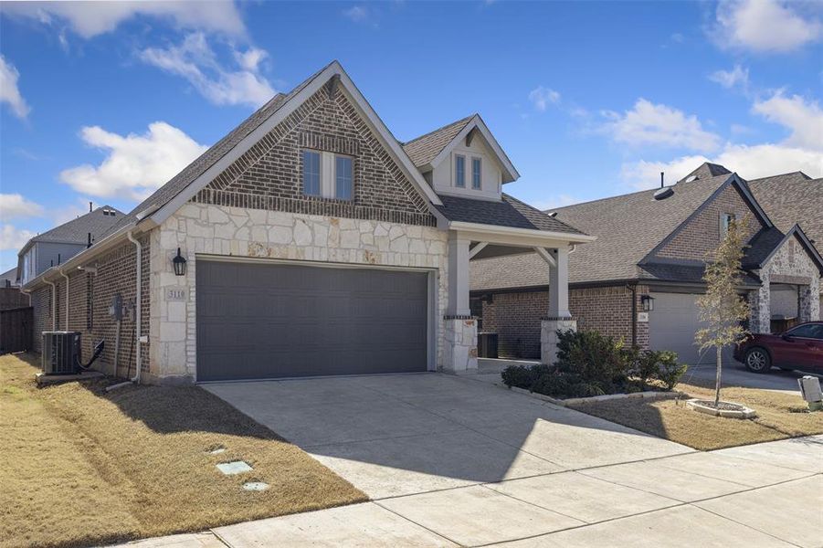 French country inspired facade with brick siding, concrete driveway, an attached garage, cooling unit, and stone siding