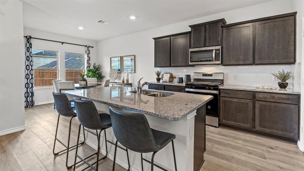 Kitchen featuring a large island with stone countertop and undermount sink, stainless steel appliances, dark wood cabinetry, and wood-finish flooring Kitchen featuring a large island with stone countertop and undermount sink, stainless steel appliances, dark wood cabinetry, and wood-finish flooring