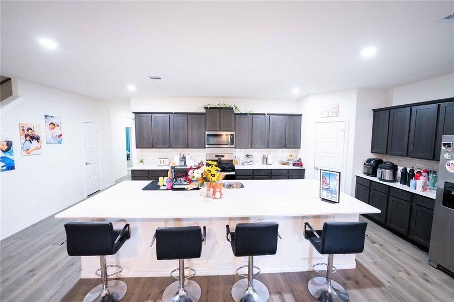 Kitchen featuring a spacious island with seating, dark wood-finish cabinetry, stainless steel appliances, and a tiled backsplash Kitchen featuring a spacious island with seating, dark wood-finish cabinetry, stainless steel appliances, and a tiled backsplash