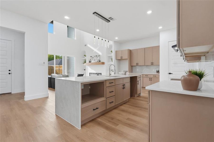 Kitchen with light wood finish cabinets, light wood-type flooring, light stone counters, an island with sink, and backsplash