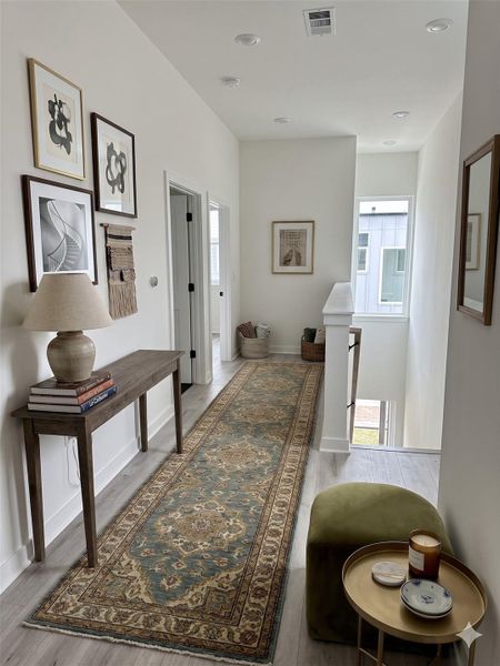 This is a bright and inviting hallway featuring a long, patterned runner rug on light wood flooring. The space is decorated with framed artwork and a console table with a lamp and books. There's also a small seating area with a side table. Natural light streams in from a window near the staircase, creating a warm atmosphere.