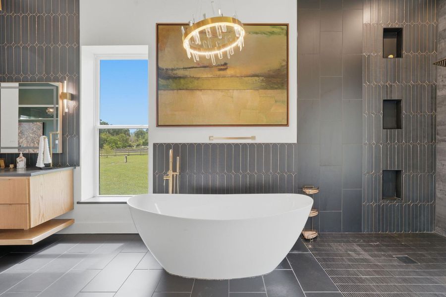 This modern bathroom features a sleek freestanding tub, elegant dark tilework, and a large window offering scenic views. A contemporary chandelier and wall art add a touch of luxury, while ample storage and stylish fixtures complete the space.