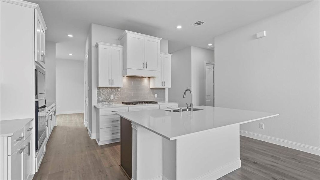 Modern kitchen featuring a large center island with integrated sink, white shaker-style cabinetry, stainless steel appliances including a built-in oven and microwave, and wood-finish flooring