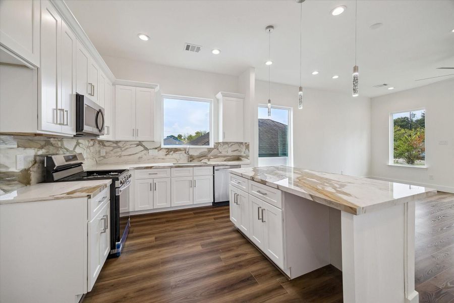 A showpiece kitchen pairs timeless white shaker cabinetry with dramatic stone-look countertops and a matching full-height backsplash. The oversized island provides generous prep space, storage, and room for casual seating, while stainless steel appliances add a polished finish. Pendant and recessed lighting brighten the space, and large windows keep the kitchen feeling fresh and open. Warm wood-look flooring ties the kitchen to the adjoining living area for a cohesive, contemporary look A showpiece kitchen pairs timeless white shaker cabinetry with dramatic stone-look countertops and a matching full-height backsplash. The oversized island provides generous prep space, storage, and room for casual seating, while stainless steel appliances add a polished finish. Pendant and recessed lighting brighten the space, and large windows keep the kitchen feeling fresh and open. Warm wood-look flooring ties the kitchen to the adjoining living area for a cohesive, contemporary look
