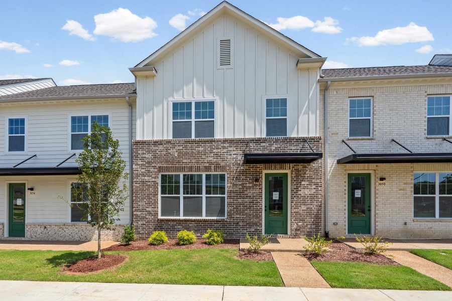 Modern townhome featuring a brick and vertical siding facade, gabled roofline, multiple windows with grilles, and a green entry door with an awning Modern townhome featuring a brick and vertical siding facade, gabled roofline, multiple windows with grilles, and a green entry door with an awning