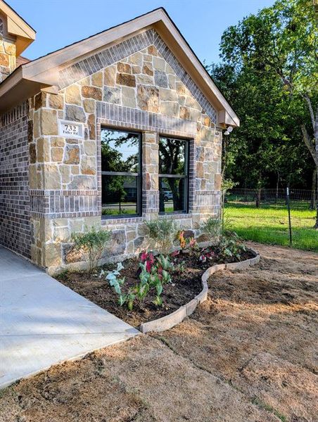 Stone and brick exterior with dark-framed windows, a landscaped planting bed, and a concrete pathway Stone and brick exterior with dark-framed windows, a landscaped planting bed, and a concrete pathway