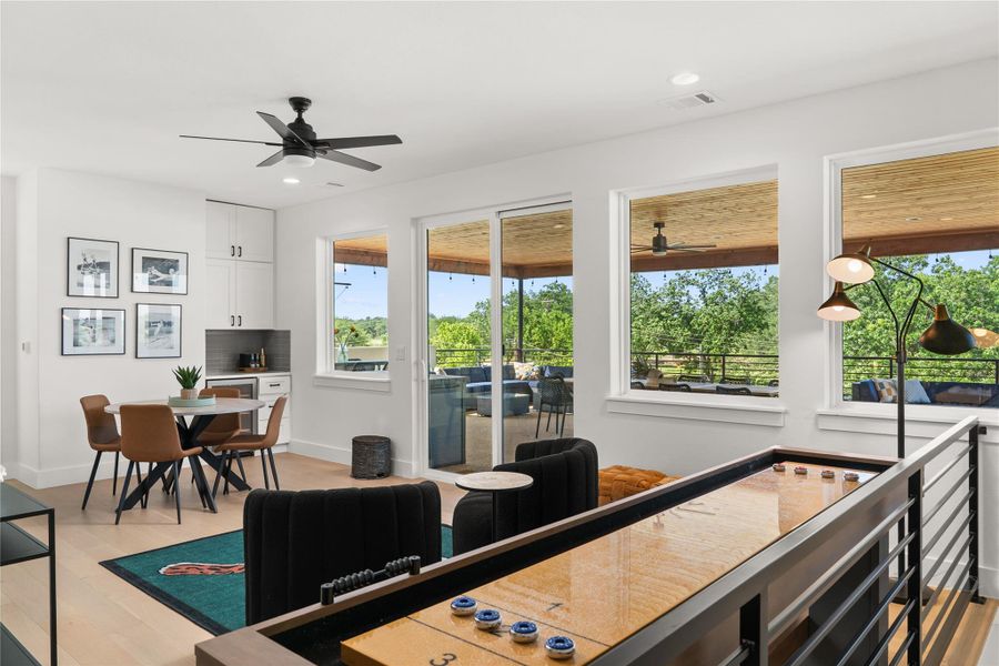 This open-concept living area features a modern design with a ceiling fan, light wood floors. Large windows and sliding doors offer plenty of natural light and lead to a spacious outdoor patio with views of greenery.
