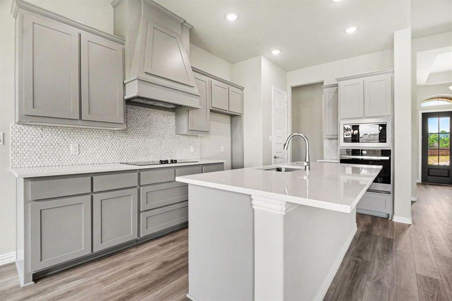 Kitchen featuring light gray cabinetry, white hexagonal tile backsplash, light-toned countertops, a central island with an undermount sink, and wood-finish flooring Kitchen featuring light gray cabinetry, white hexagonal tile backsplash, light-toned countertops, a central island with an undermount sink, and wood-finish flooring