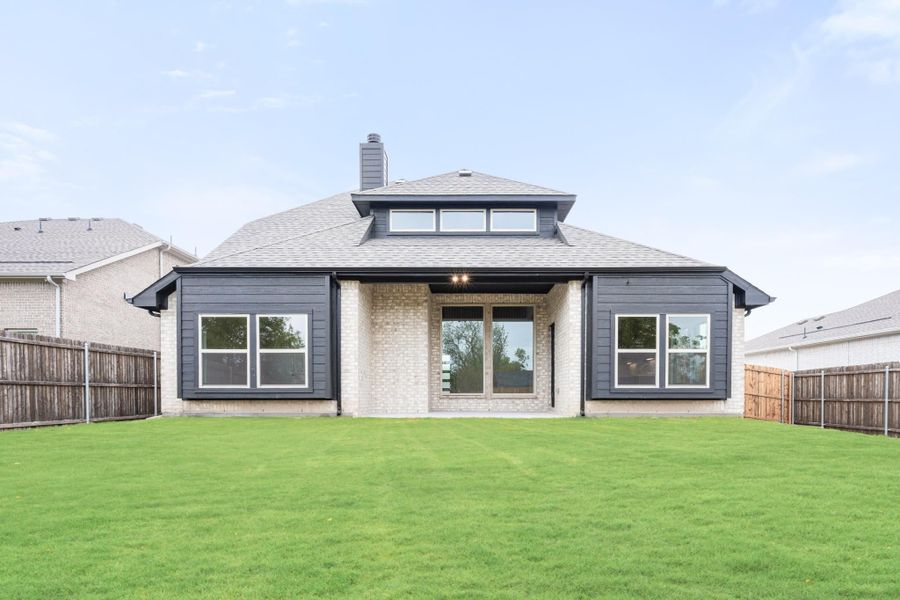 Rear exterior of single-story home with dark siding, brick accents, large windows, and fenced backyard with green lawn.