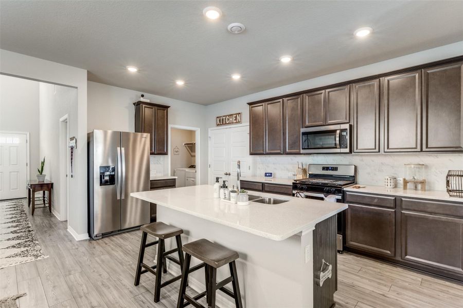 Kitchen featuring dark brown cabinetry, stainless steel appliances, a kitchen bar, decorative backsplash, and light stone countertops