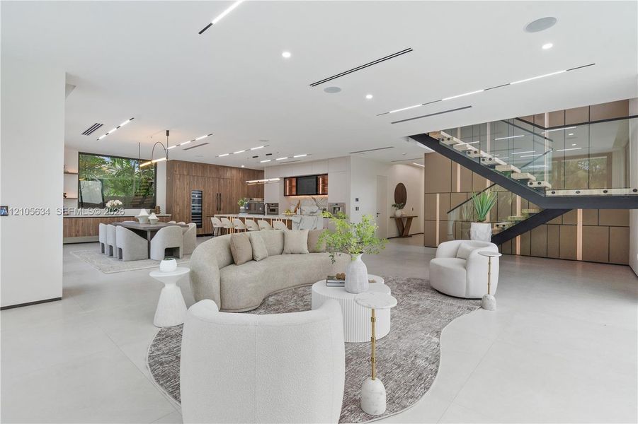 Open-concept floor plan featuring expansive stone-tile flooring, recessed linear lighting, custom wood-paneled cabinetry, and a floating staircase with glass railings