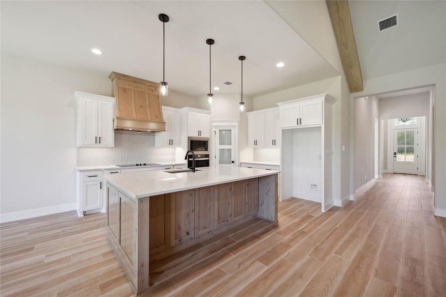 Kitchen with decorative backsplash, light wood-type flooring, premium range hood, a center island with sink, and white cabinetry