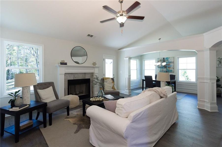 Living room with dark wood-type flooring, decorative columns, lofted ceiling, ceiling fan with notable chandelier, and a tile fireplace