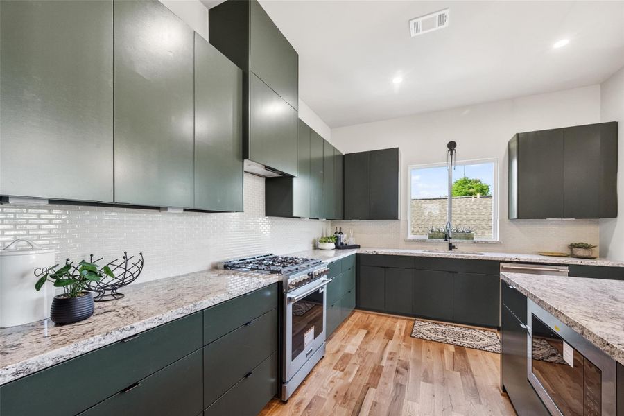 Natural light from the sink-side window floods the kitchen, showing off the clean geometry of the flat-front cabinetry. The stone countertops wrap the full perimeter of the cooking wall, providing generous prep space throughout.