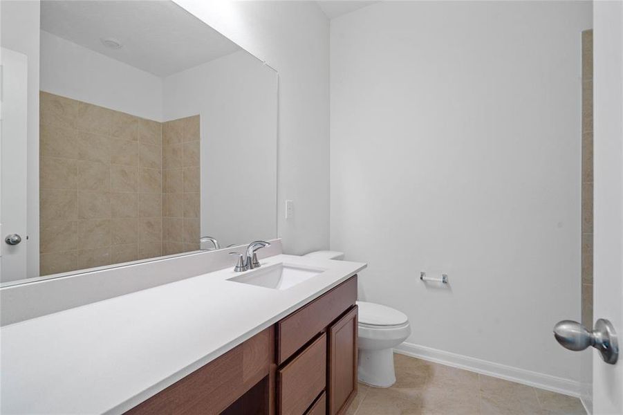 Secondary bath features tile flooring, bath/shower combo with tile surround, stained wood cabinets, beautiful light countertops, mirror, dark, sleek fixtures and modern finishes.