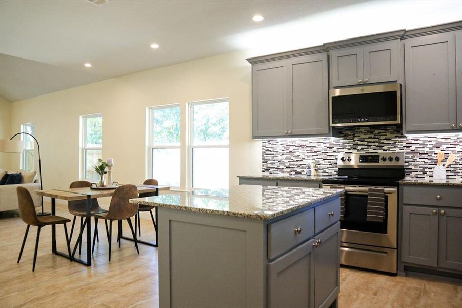 Kitchen with light stone countertops, a kitchen island, stainless steel appliances, gray cabinets, and decorative backsplash