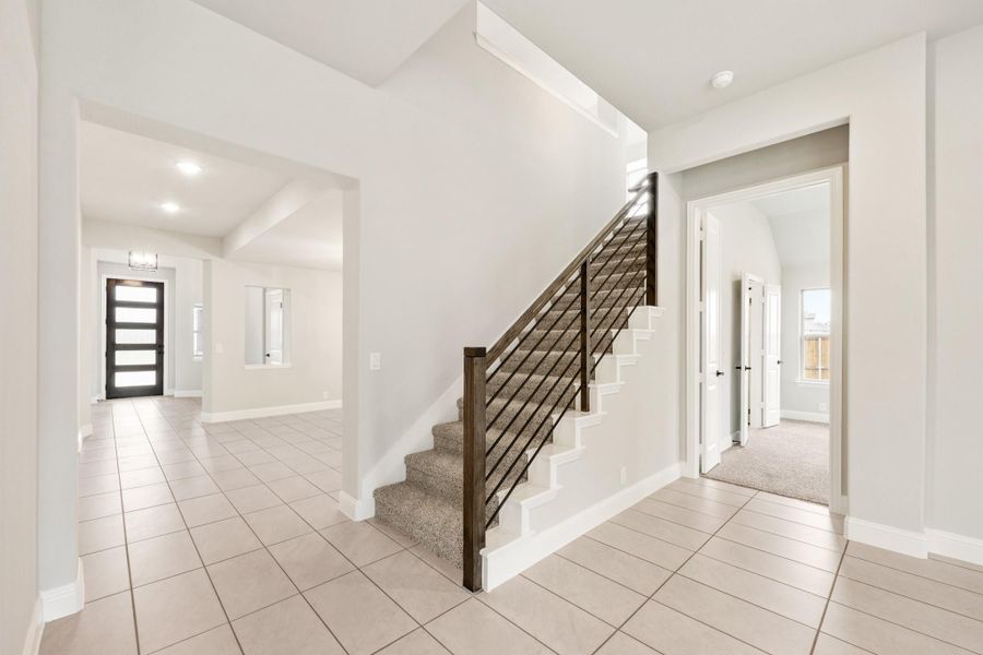 Open foyer with carpeted staircase, dark metal railing, tile floors, and modern front door visible in background Open foyer with carpeted staircase, dark metal railing, tile floors, and modern front door visible in background