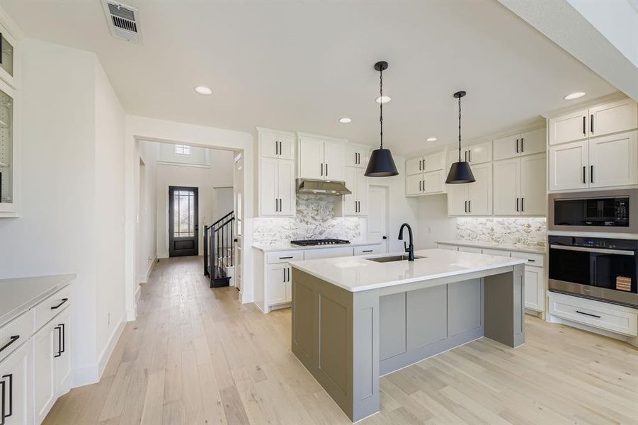 Kitchen featuring stainless steel appliances, light wood-style flooring, light countertops, a center island with sink, and decorative light fixtures Kitchen featuring stainless steel appliances, light wood-style flooring, light countertops, a center island with sink, and decorative light fixtures