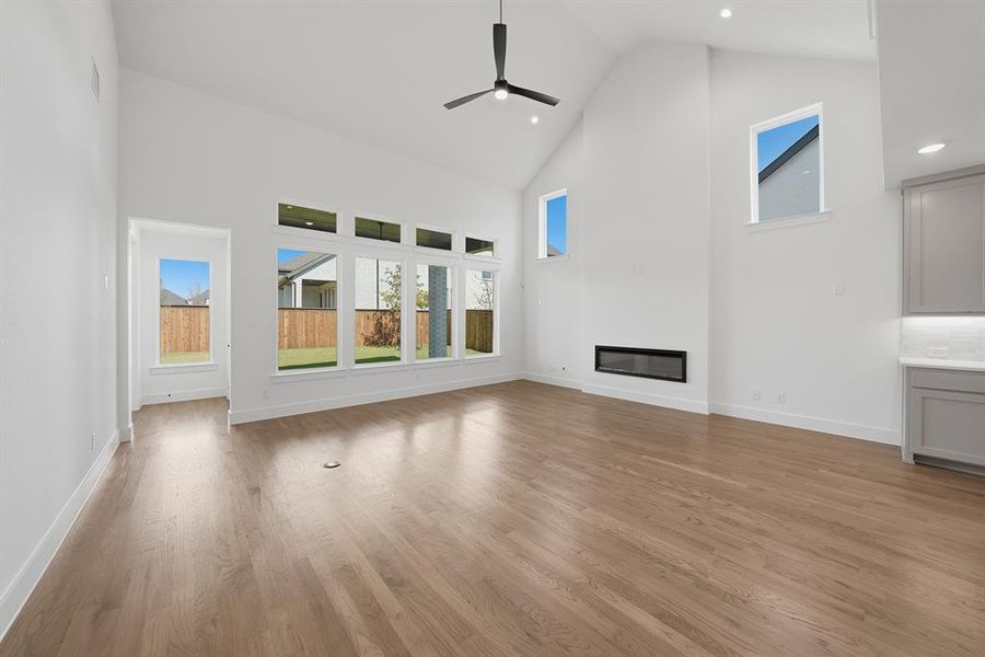 Unfurnished living room with high vaulted ceiling, a glass covered fireplace, light wood-type flooring, a ceiling fan, and recessed lighting Unfurnished living room with high vaulted ceiling, a glass covered fireplace, light wood-type flooring, a ceiling fan, and recessed lighting