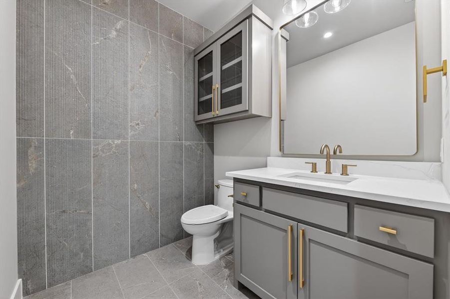Powder room featuring full-height tiled walls, tiled flooring, a gray vanity with gold-tone hardware, a white countertop, and a rectangular mirror with a gold-tone frame Powder room featuring full-height tiled walls, tiled flooring, a gray vanity with gold-tone hardware, a white countertop, and a rectangular mirror with a gold-tone frame