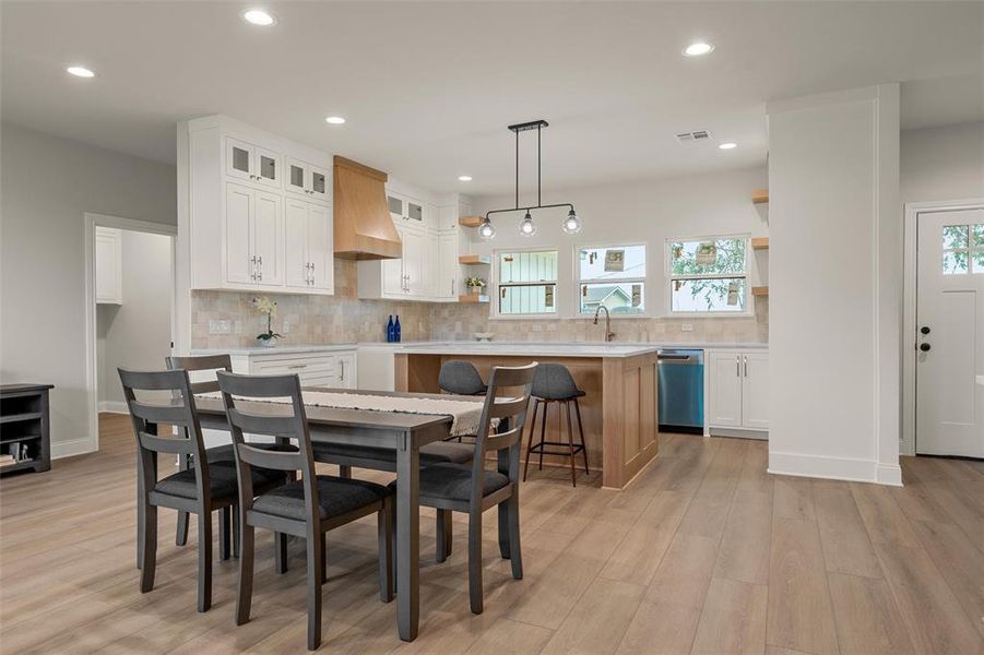 Bright kitchen and dining area featuring light wood-finish flooring, white cabinetry, a wood range hood, a tiled backsplash, and a central island with bar seating