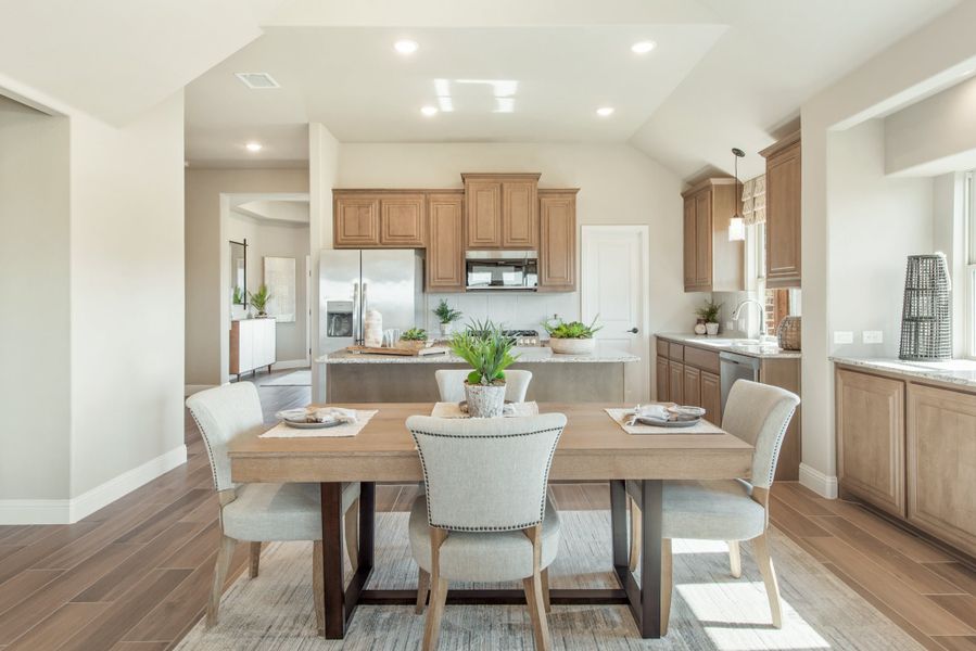 Open dining area with wood table and upholstered chairs, adjacent to kitchen with tan cabinets and stainless appliances