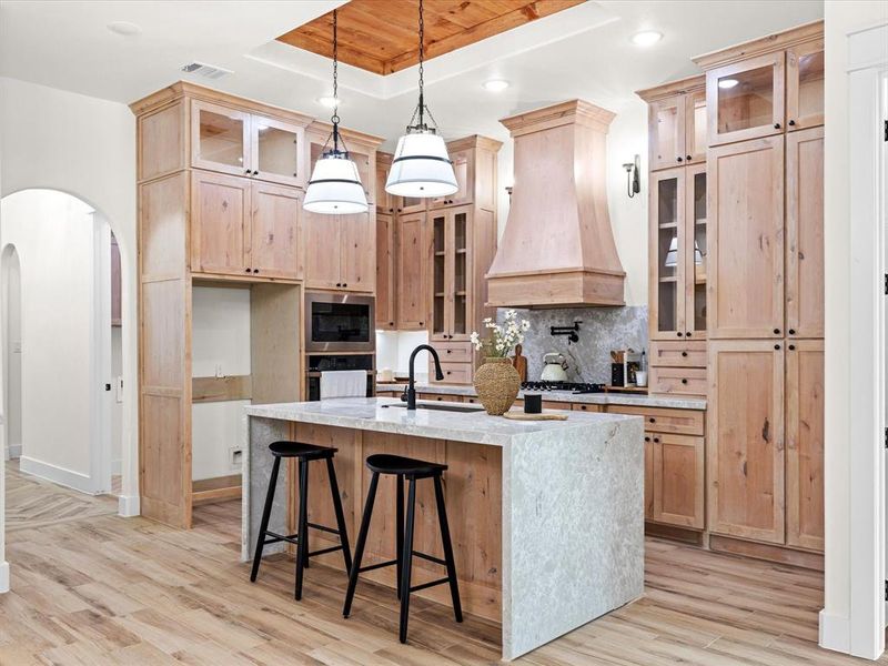 Kitchen with glass insert cabinets, a raised ceiling, light stone countertops, a kitchen bar, and light brown cabinets