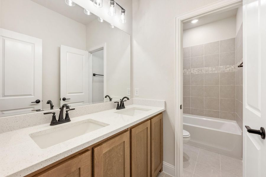 Bathroom with double sink vanity, wood cabinets, and tiled tub-shower combo visible through doorway