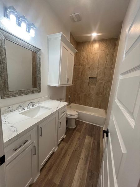 This bathroom features a sleek design with medium gray/ wood water proof flooring, a white vanity with a marble countertop, and ample storage. The shower-tub combo is accented with warm, textured tiles, and bright lighting enhances the space.