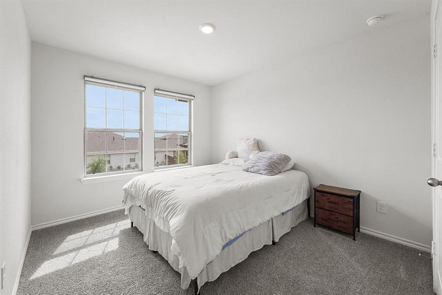 Bright bedroom featuring two large windows with roller shades, neutral gray carpeting, white painted walls, a recessed ceiling light, and white baseboards