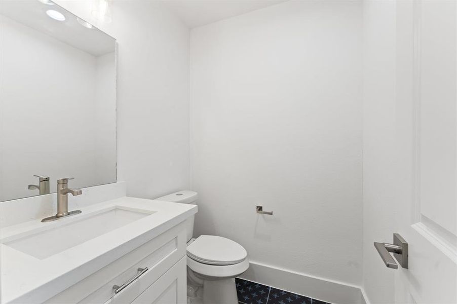 This is a modern, compact bathroom with white walls and a sleek design. It features a white vanity with a contemporary faucet, a large mirror, and a toilet. The dark, patterned floor adds a touch of style to the bright space.