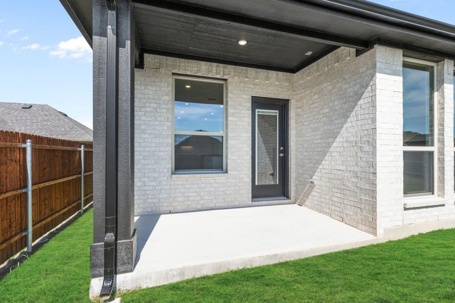 Covered back patio with concrete slab, gray brick exterior, dark entry door, and green lawn Covered back patio with concrete slab, gray brick exterior, dark entry door, and green lawn