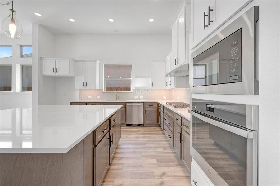 Spacious kitchen featuring a large central island with light-toned countertops, white upper cabinetry, wood-finish lower cabinetry, stainless steel appliances, and wood-finish flooring Spacious kitchen featuring a large central island with light-toned countertops, white upper cabinetry, wood-finish lower cabinetry, stainless steel appliances, and wood-finish flooring