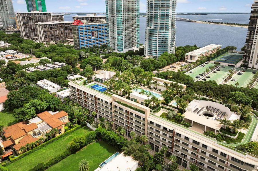 Aerial perspective of a multi-story residential complex featuring a rooftop swimming pool, a blue-surfaced tennis court, a circular courtyard area, and landscaped grounds Aerial perspective of a multi-story residential complex featuring a rooftop swimming pool, a blue-surfaced tennis court, a circular courtyard area, and landscaped grounds