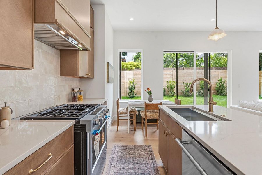 Gourmet kitchen featuring a gas range with a stainless steel vent hood, white countertops, wood-finish cabinetry, a large kitchen island with an undermount sink and brass faucet, and multiple large windows overlooking a private yard